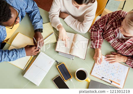 Top view shot of diverse team of colleagues planning monthly schedule discussing agenda while sitting at meeting table, cluttered with notebooks and office stationery at workplace Top view shot of diverse team of colleagues planning monthly schedule discussing agenda while sitting at meeting table, cluttered with notebooks and office stationery at workplace 121301219