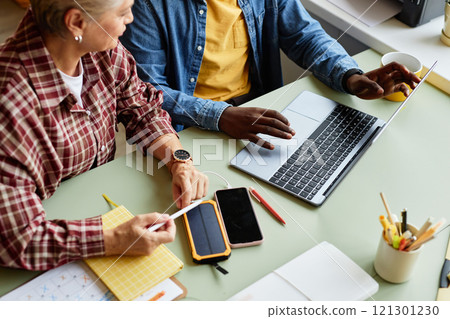 Cropped shot of African American man collaborating with senior colleague using laptop pointing at screen, during work meeting at desk cluttered with phones and office stationery 121301230