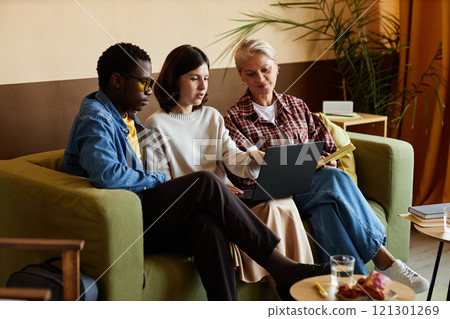 Multigenerational team of office workers discussing project details pointing at laptop screen while sitting on couch at workplace 121301269