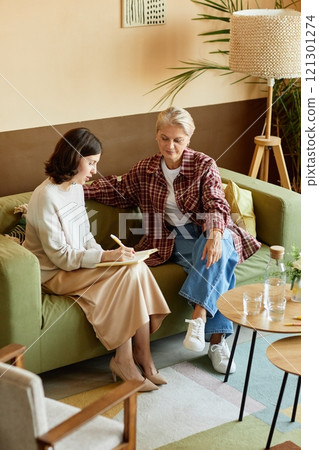 Vertical shot of young woman consulting with experienced senior colleague dressed in casual yet elegant outfit, while writing down records in notebook sitting on couch at cozy office workplace Vertical shot of young woman consulting with experienced senior colleague dressed in casual yet elegant outfit, while writing down records in notebook sitting on couch at cozy office workplace 121301274