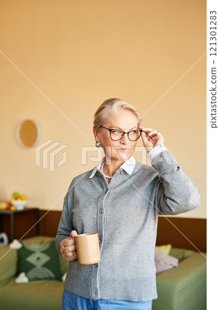Medium shot of stylish mature woman looking over glasses with confident look holding cup of coffee while posing in modern cozy office with beige walls, copy space 121301283