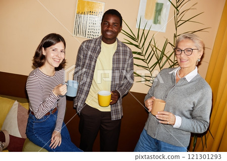 Portrait shot of senior and younger colleagues from different ethnic backgrounds smiling at camera while having coffee break in cozy office, camera flash Portrait shot of senior and younger colleagues from different ethnic backgrounds smiling at camera while having coffee break in cozy office, camera flash 121301293