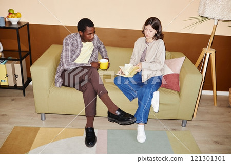 Full length shot of African American man and young woman reading records in notebook during working meeting leisurely sitting on comfortable couch in modern office, camera flash Full length shot of African American man and young woman reading records in notebook during working meeting leisurely sitting on comfortable couch in modern office, camera flash 121301301