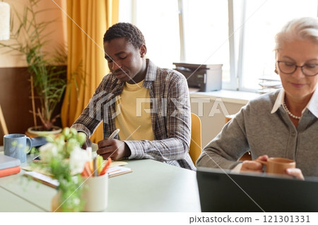 Medium shot of young African American corporate worker concentrated on writing notes on paper sitting at desk in cozy modern office with beige color scheme, copy space Medium shot of young African American corporate worker concentrated on writing notes on paper sitting at desk in cozy modern office with beige color scheme, copy space 121301331