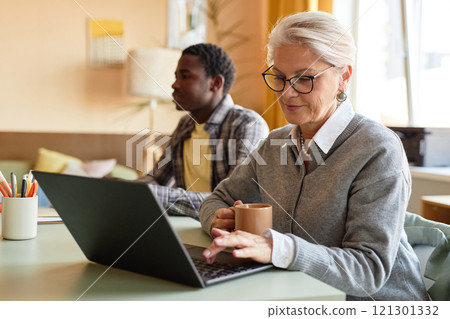 Medium shot of mature businesswoman wearing elegant glasses typing on laptop keyboard while writing business report sitting at desk in corporate office, copy space Medium shot of mature businesswoman wearing elegant glasses typing on laptop keyboard while writing business report sitting at desk in corporate office, copy space 121301332