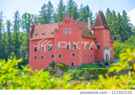 Cervena Lhota Castle, with its distinctive red facade, is set on a rocky island in a serene lake, surrounded by lush greenery and tall trees, creating a picturesque view in Czechia. Cervena Lhota Castle, with its distinctive red facade, is set on a rocky island in a serene lake, surrounded by lush greenery and tall trees, creating a picturesque view in Czechia. 121302529