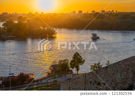 The sun sets over the Sava River, casting golden hues across the water. From Kalamegdan fortress in Belgrade, the view showcases peaceful nature and city skyline blending beautifully. The sun sets over the Sava River, casting golden hues across the water. From Kalamegdan fortress in Belgrade, the view showcases peaceful nature and city skyline blending beautifully. 121302556