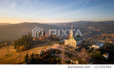 Beneath a golden autumn sunset, the Tanvaldsky Spicak lookout tower stands tall in the Jizera Mountains, surrounded by colorful foliage, inviting exploration and admiration. 121302568