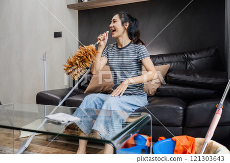Asian woman happily use feather pick as microphone while cleaning house on the weekend Asian woman happily use feather pick as microphone while cleaning house on the weekend 121303643