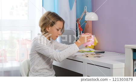 Young girl sits at her table in her room shaking coins from a pink piggy bank as she checks her savings. She explores the importance of personal finance and money management Young girl sits at her table in her room shaking coins from a pink piggy bank as she checks her savings. She explores the importance of personal finance and money management 121303879