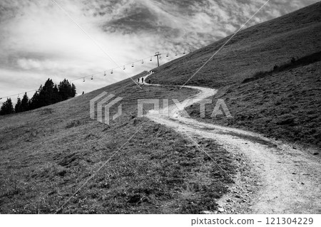 A winding road ascends through grassy hills under a cloudy sky. Hikers can be seen making their way toward the mountain summit, surrounded by lush greenery. 121304229