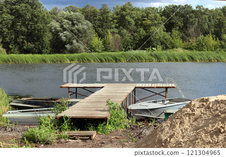 Old boats docked at a rustic village pier alongside a tranquil river surrounded by lush greenery 121304965