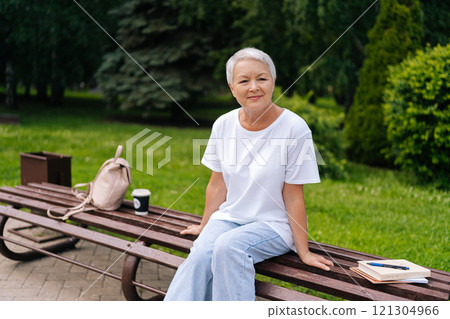 Thoughtful elderly woman with silver hair relaxing on park bench with backpack, books, and cup of coffee, savoring tranquility. Serene European gray-haired older female enjoying moment of relaxation. Thoughtful elderly woman with silver hair relaxing on park bench with backpack, books, and cup of coffee, savoring tranquility. Serene European gray-haired older female enjoying moment of relaxation. 121304966