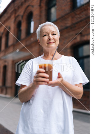 Vertical portrait of older woman drinking iced coffee outdoors, smiling looking at camera in urban setting with brick building. European gray-haired older female enjoying free time outdoors. Vertical portrait of older woman drinking iced coffee outdoors, smiling looking at camera in urban setting with brick building. European gray-haired older female enjoying free time outdoors. 121304972