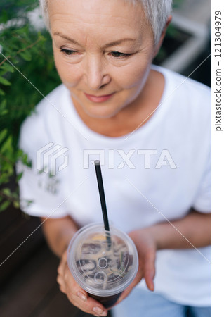 Vertical top view shot of senior woman drinking refreshing iced coffee sitting at outdoors cafe, relishing peaceful moment in bustling city on summer day, enjoying free time in retirement Vertical top view shot of senior woman drinking refreshing iced coffee sitting at outdoors cafe, relishing peaceful moment in bustling city on summer day, enjoying free time in retirement 121304979