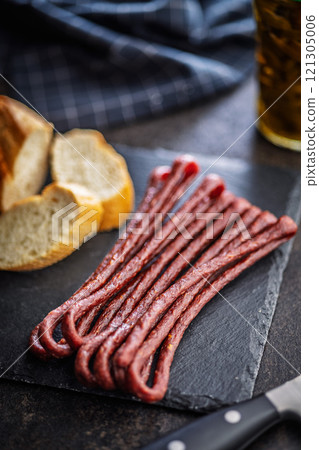 Smoked kabanos. Mini dried thin sausages on cutting board on black table. 121305006