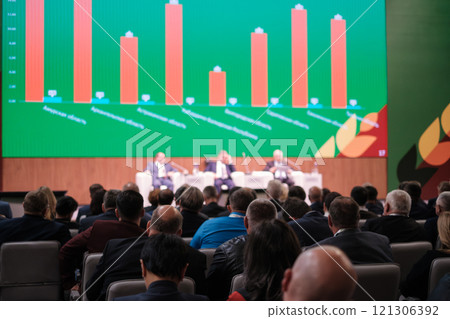 Conference attendees watching panelists discussing data on a large screen 121306392