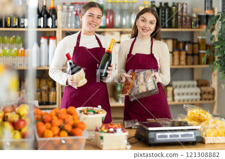 Two sales girls pose behind a store counter with groceries, sweets and wine in their hands Two sales girls pose behind a store counter with groceries, sweets and wine in their hands 121308882