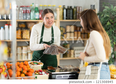 Woman store employee offers customer to choose sweets, shows packed various dessert options. Woman store employee offers customer to choose sweets, shows packed various dessert options. 121308945