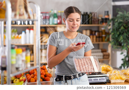 Girl shopper scanning QR code on raw sausages label in grocery department of supermarket Girl shopper scanning QR code on raw sausages label in grocery department of supermarket 121309369