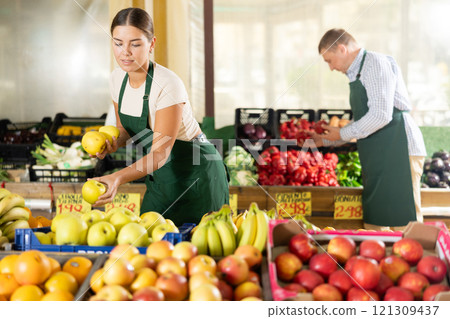 Girl seller of garden-stuff store works near showcase, sorting apples, checking goods 121309437