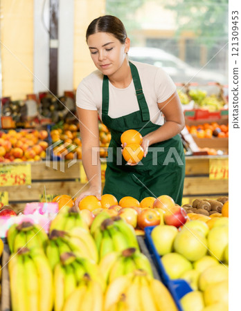 Interested salesgirl laying out oranges on display stand in supermarket Interested salesgirl laying out oranges on display stand in supermarket 121309453