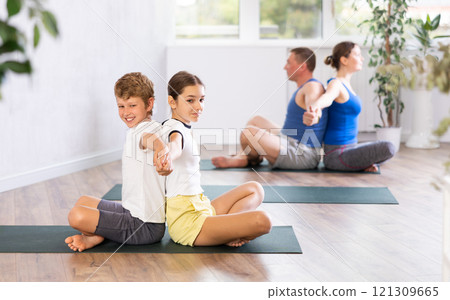School children and their parents sitting back to back on mats during family yoga training together in gym fitness center indoors School children and their parents sitting back to back on mats during family yoga training together in gym fitness center indoors 121309665