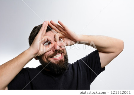 Playful man makes binoculars with hands, looking through with wide smile. 121312061