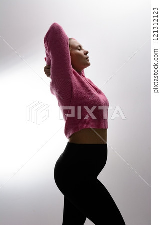 Pregnant woman in pink sweater and black leggings, arms raised, against a white background. 121312123