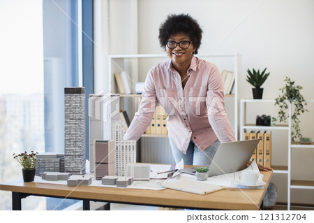 African American woman architect in 30s develops project with skyscraper model on desk, wearing glasses. African American woman architect in 30s develops project with skyscraper model on desk, wearing glasses. 121312704