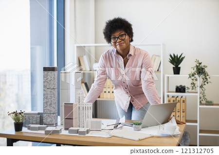 African American woman architect in 30s develops project with skyscraper model on desk, wearing glasses. African American woman architect in 30s develops project with skyscraper model on desk, wearing glasses. 121312705