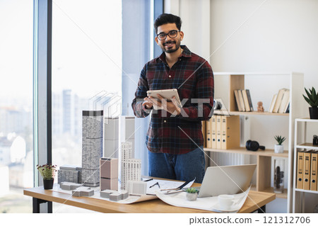 Indian male architect develops modern city project using digital tablet in creative workspace. Tall building models on desk symbolize innovative urban design. Age group young adult to middle-aged. Indian male architect develops modern city project using digital tablet in creative workspace. Tall building models on desk symbolize innovative urban design. Age group young adult to middle-aged. 121312706