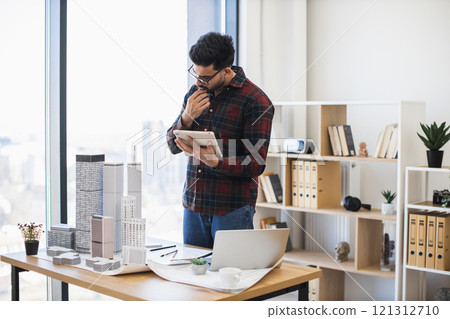 Indian male architect in modern office using digital tablet to develop city project with model buildings, emphasizing creativity and concentration. Indian male architect in modern office using digital tablet to develop city project with model buildings, emphasizing creativity and concentration. 121312710