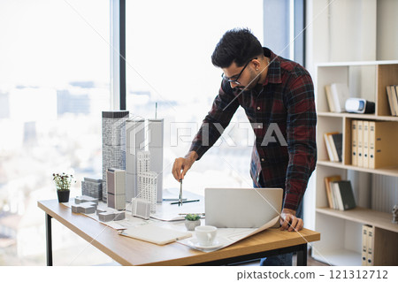 Indian male architect in flannel shirt measuring dimensions of skyscraper model with compass, developing city design project in modern office setting. 121312712