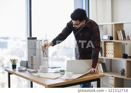 Indian male architect in twenties develops urban project using building models and compass. He stands at desk in modern office, focusing on precision and innovation in architectural design. Indian male architect in twenties develops urban project using building models and compass. He stands at desk in modern office, focusing on precision and innovation in architectural design. 121312718