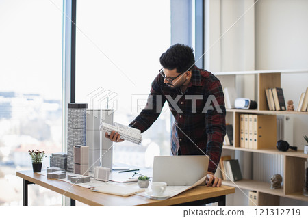 Indian male architect focuses on creating detailed city model for modern urban development, showcasing creativity and precision in office environment with natural light. 121312719