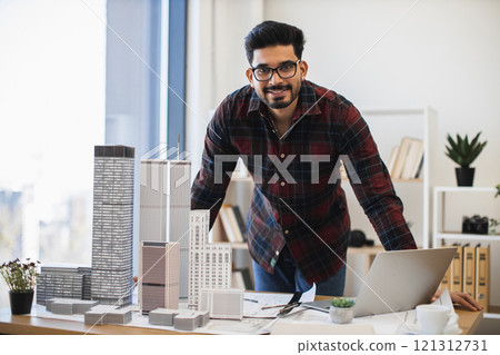 Indian male architect in 30s designing city architecture model at office desk with laptop and buildings. Creative professional working on urban development project in modern workspace. Indian male architect in 30s designing city architecture model at office desk with laptop and buildings. Creative professional working on urban development project in modern workspace. 121312731