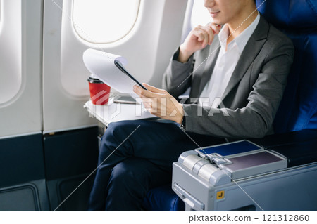 Asian businessman or entrepreneur in a formal suit on an airplane sitting in business class using a phone, computer laptop 121312860