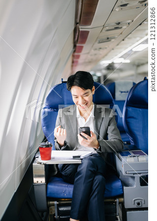 Asian businessman or entrepreneur in a formal suit on an airplane sitting in business class using a phone, computer laptop 121312866