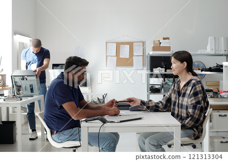 People collaborating in modern office setting with electronic gadgets. Man and woman exchanging devices, while another person in background working with technology People collaborating in modern office setting with electronic gadgets. Man and woman exchanging devices, while another person in background working with technology 121313304