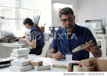 Bearded middle-aged man engrossed in examining packaging box at his desk in contemporary office setting with colleague working diligently in background Bearded middle-aged man engrossed in examining packaging box at his desk in contemporary office setting with colleague working diligently in background 121313331