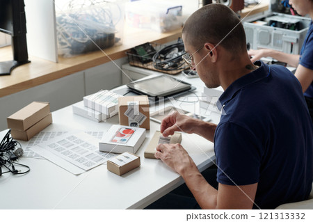 Man seated at desk organizing cardboard boxes and office supplies, preparing for project or shipment. Background includes office equipment and shelves with storage containers 121313332