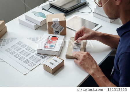Person seen packaging and labeling products at organized workspace with various boxes, labels, and a tablet visible on table, appearing focused on work process Person seen packaging and labeling products at organized workspace with various boxes, labels, and a tablet visible on table, appearing focused on work process 121313333