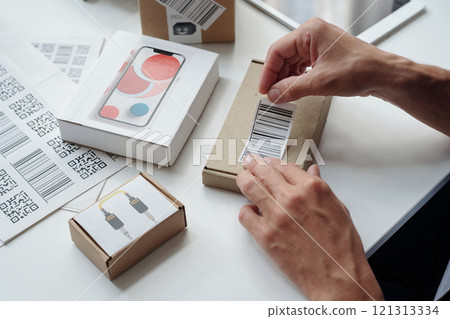 Hands labeling cardboard boxes with QR codes in warehouse setting. Multiple packages visible, along with a phone box and charging cables, creating an organized workspace Hands labeling cardboard boxes with QR codes in warehouse setting. Multiple packages visible, along with a phone box and charging cables, creating an organized workspace 121313334