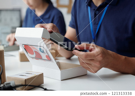 Close-up of hands inspecting and testing mobile device at workplace showing technical workflow without visible faces. Colleagues in background contributing to process Close-up of hands inspecting and testing mobile device at workplace showing technical workflow without visible faces. Colleagues in background contributing to process 121313338