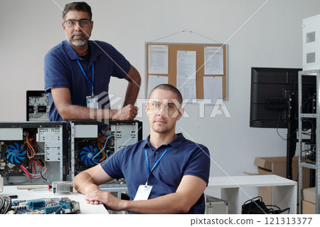 Portrait of two computer technicians wearing blue shirts, standing near disassembled computer units, looking thoughtfully at camera, surrounded by various electronic components 121313377