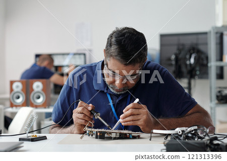 Portrait of man concentrating on soldering circuits at a workbench in an electronics lab surrounded by equipment and tools actively engaging in detailed precision work 121313396