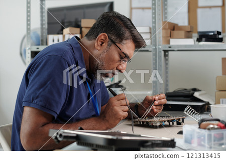 Man concentrating on fixing circuit board at workspace featuring shelves filled with boxes and equipment. Precision in handling small electronic components visible in workspace 121313415
