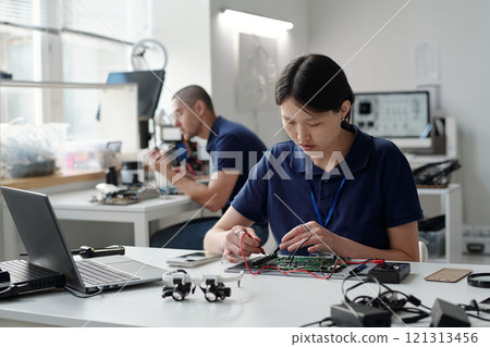 Engineers wearing blue shirts working on circuit boards in modern office setting. People focusing on intricate wiring and device assembly, surrounded by various electronic gadgets and tools Engineers wearing blue shirts working on circuit boards in modern office setting. People focusing on intricate wiring and device assembly, surrounded by various electronic gadgets and tools 121313456