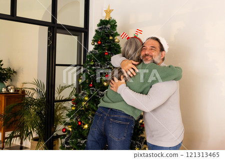 Mature couple hugging joyfully by Christmas tree at home, celebrating holiday spirit 121313465